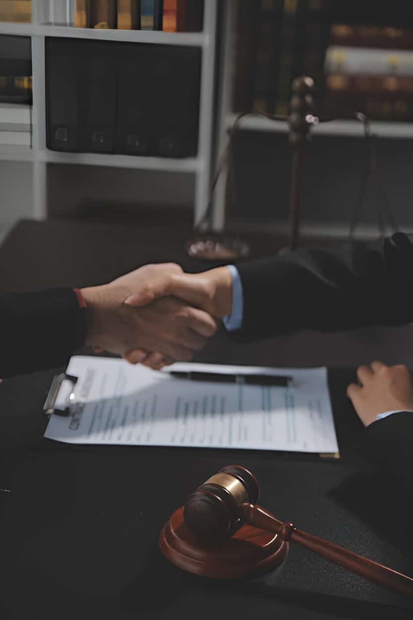 dispute-resolution Two people shaking hands over a desk with a legal document, gavel, and scales of justice in a law office setting.