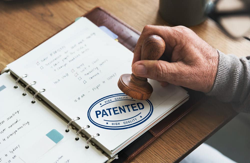 patents A hand stamping 'PATENTED HIGH QUALITY' on a notebook with handwritten notes, placed on a wooden desk with a leather folder and a cup in the background.